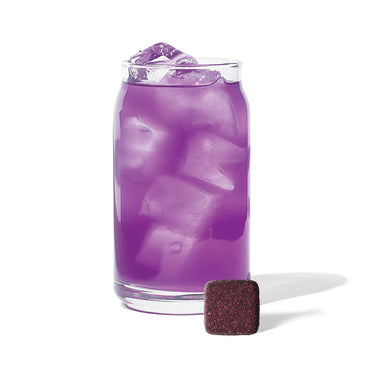 A clear glass with ice and a vibrant purple drink sits on a white background. In front is a small, dark purple cube of 💜 Ube Tea Drops® – A Sweet, Nutty, & Creamy Purple Delight! 🍠✨, perfect for making purple yam tea.